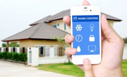 hand holding white mobile smart phone with smart home application on the screen over blurred house background, smart home concept